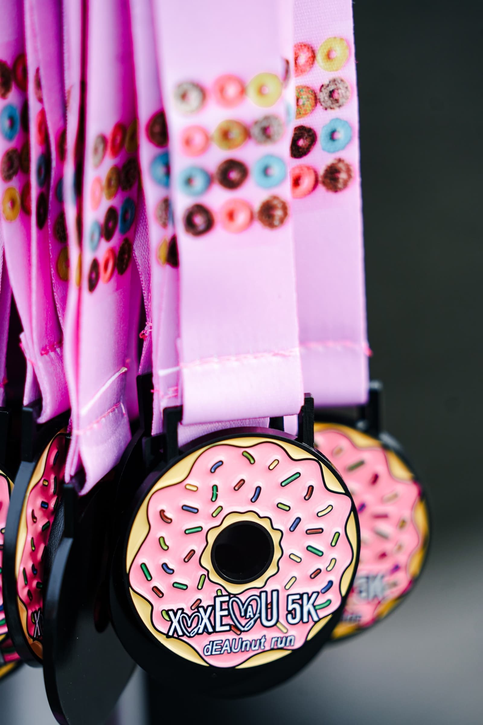 A close-up of several pink medals featuring a donut design with colorful sprinkles and "XoXo 5K DEANut run" text, hanging from pink lanyards decorated with tiny donut images.