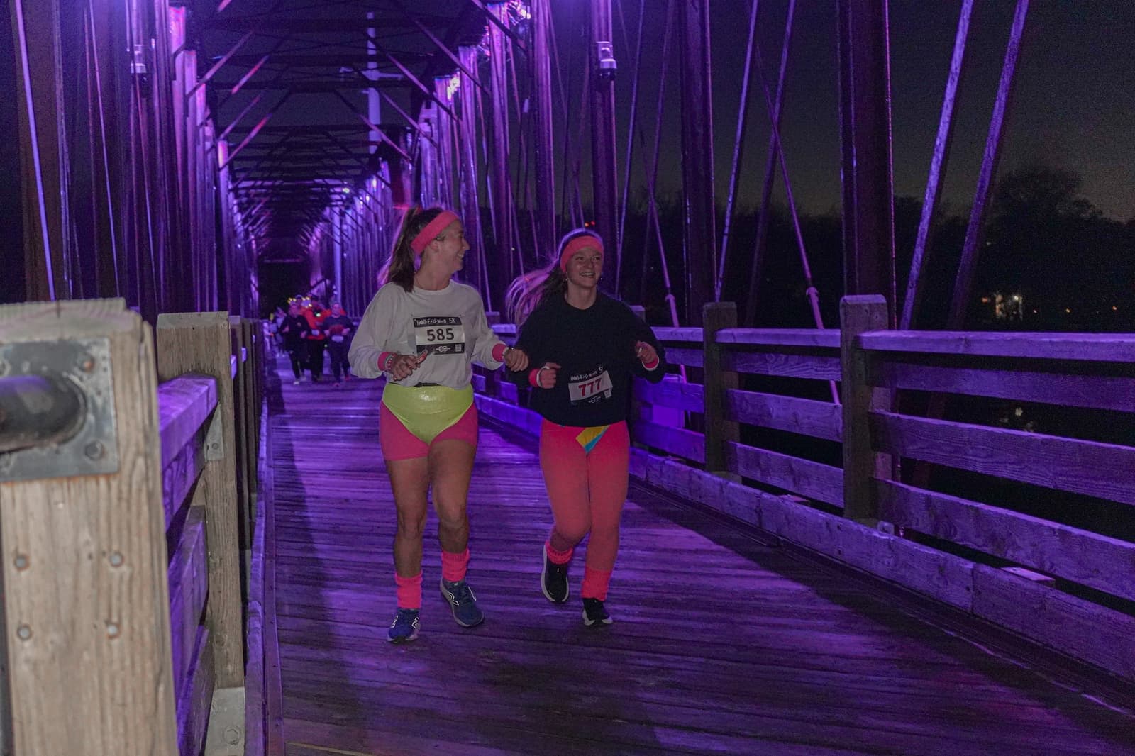 Two women in neon workout clothes and numbered race bibs run and smile together on a wooden bridge at night, lit by purple lights. Other runners are visible in the background.