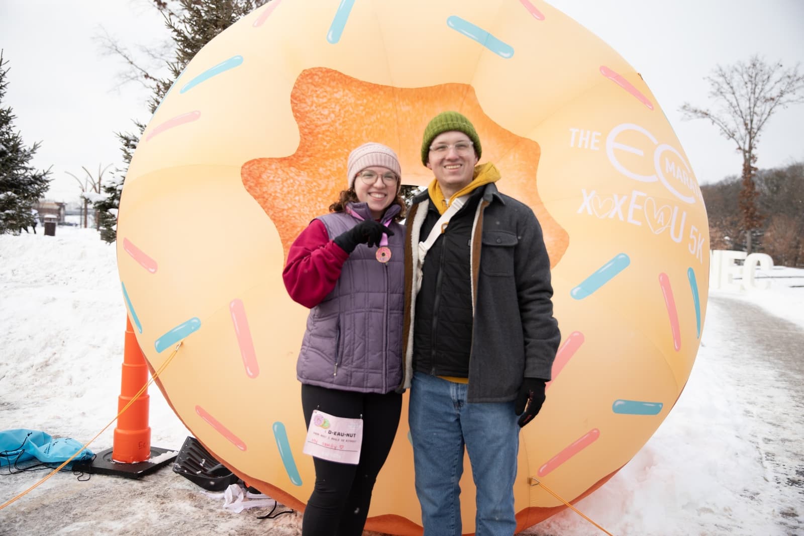 Two people smiling and posing in winter clothes in front of a large inflatable donut outdoors on a snowy day. One holds a medal and a race bib, suggesting they participated in an event.
