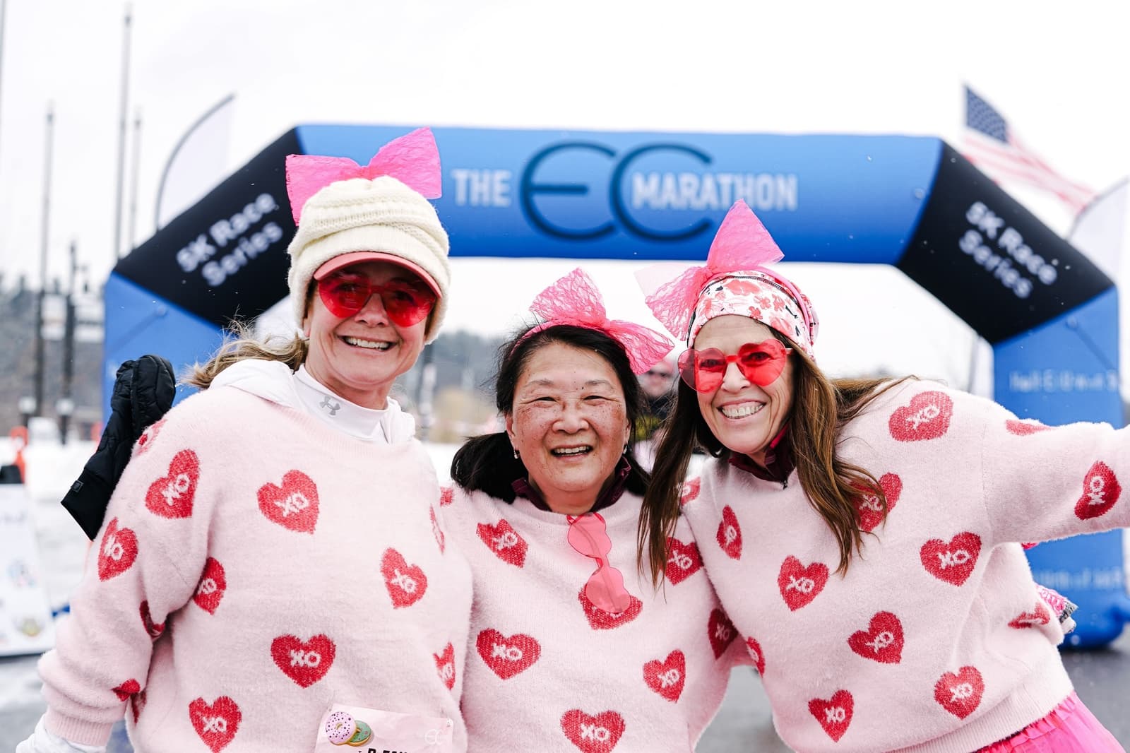 Three women in matching pink sweaters with red hearts and "XO" smile in front of a marathon finish line. They wear pink bows, festive glasses, and stand arm in arm, celebrating together outdoors.