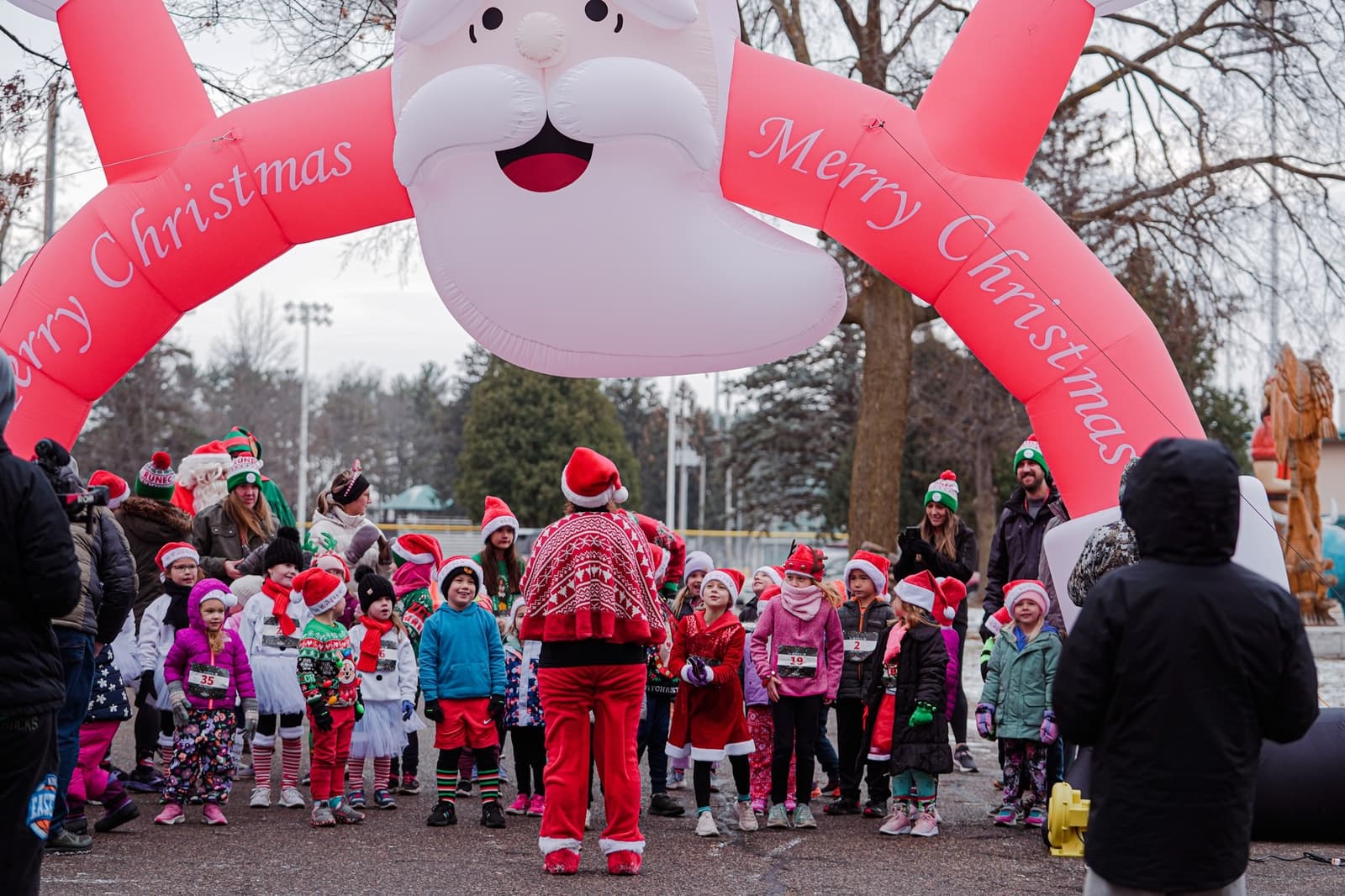 A group of children in festive clothing and Santa hats gather under a large inflatable Santa arch that says "Merry Christmas," while adults stand nearby at an outdoor holiday event.