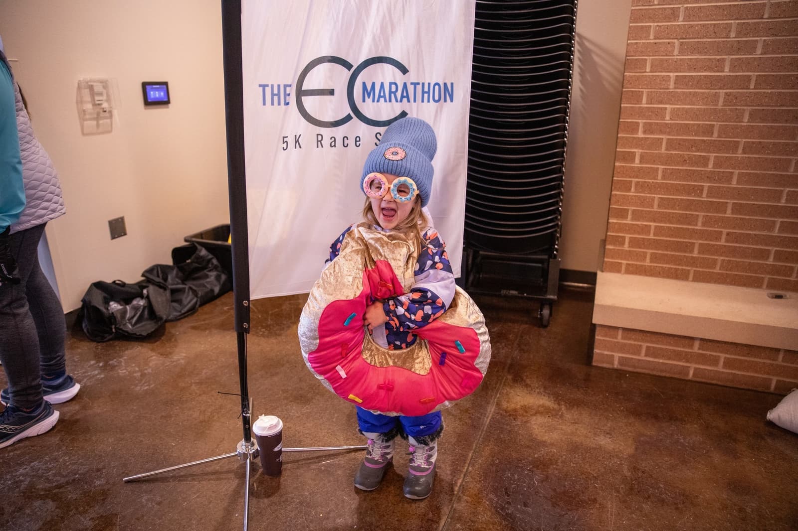 A young child wearing winter clothes, large glasses, a knit hat, and a donut costume smiles in front of a "EC Marathon 5K Race" banner indoors. A cup of hot chocolate is on the floor nearby.