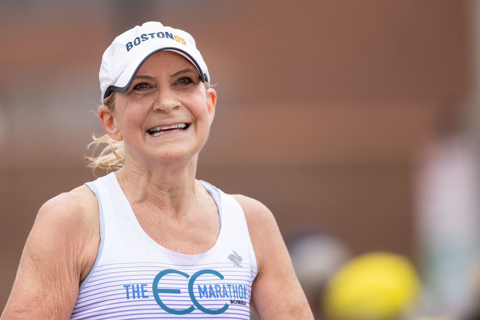 A woman wearing a white cap with "Boston 26.2" and a white tank top with blue text smiles while running outside. The background is blurred, focusing on her expression and attire.