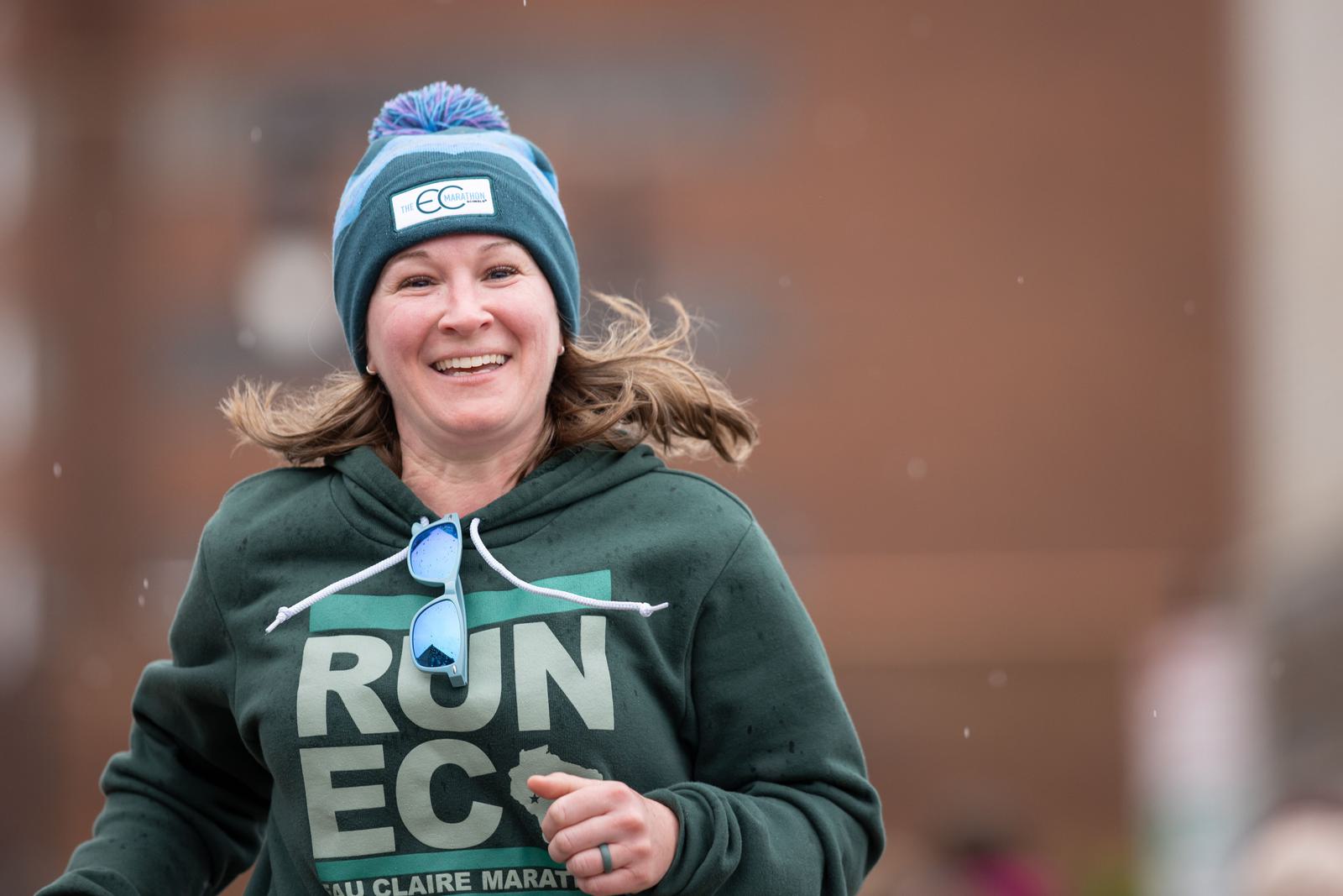 A person runs outdoors wearing a green hoodie and a blue beanie with "EC" on it. They are smiling, with sunglasses hanging from the hoodie. The background is blurred, suggesting movement.