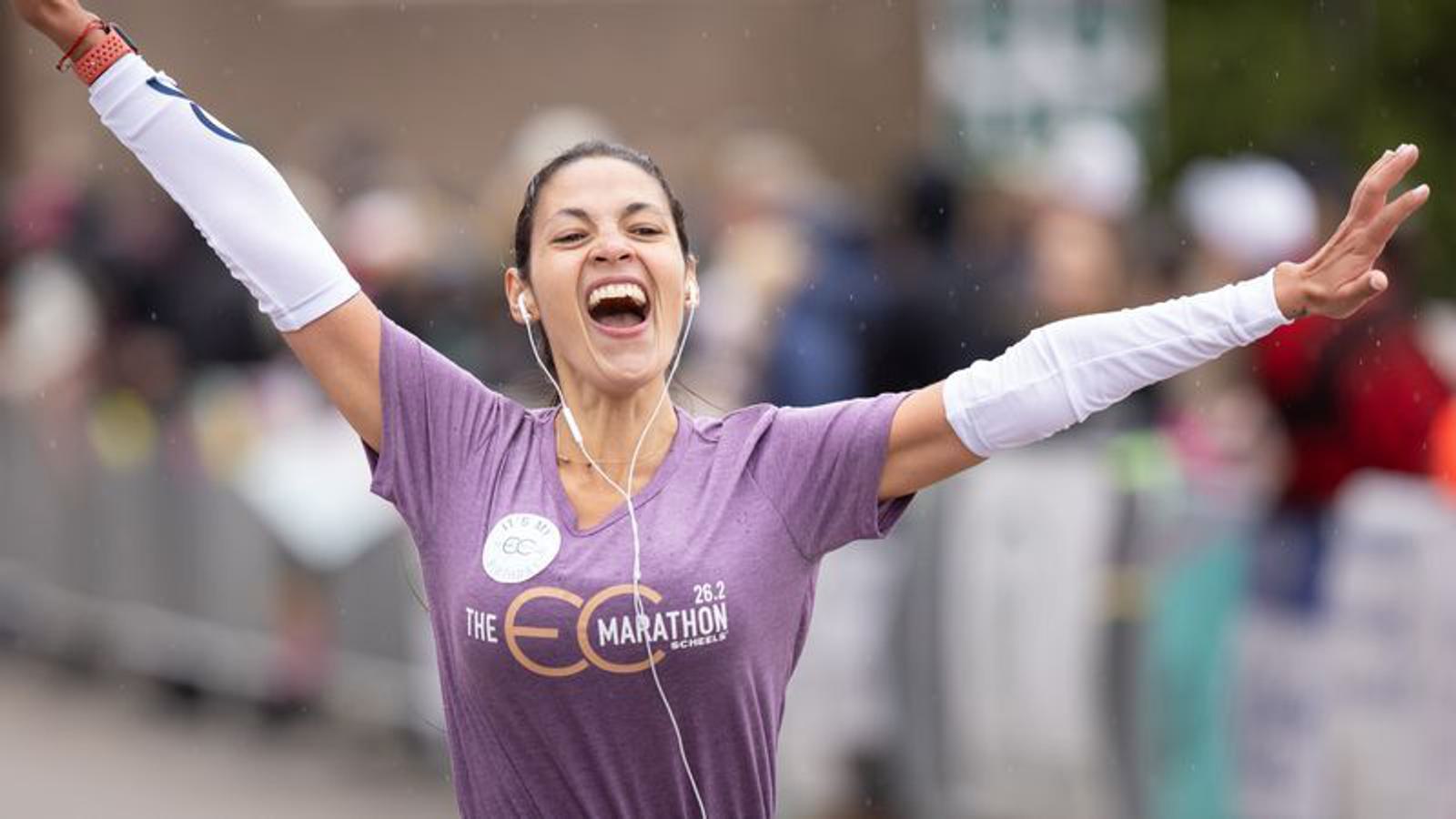 A woman wearing a purple shirt with "The Eco Marathon" on it crosses the finish line, arms raised in triumph. She has earbuds in and is smiling widely, expressing joy. The background is blurred, featuring spectators.