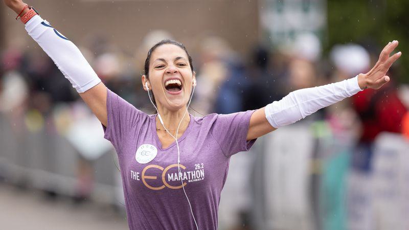 A woman wearing a purple shirt with "The Eco Marathon" on it crosses the finish line, arms raised in triumph. She has earbuds in and is smiling widely, expressing joy. The background is blurred, featuring spectators.