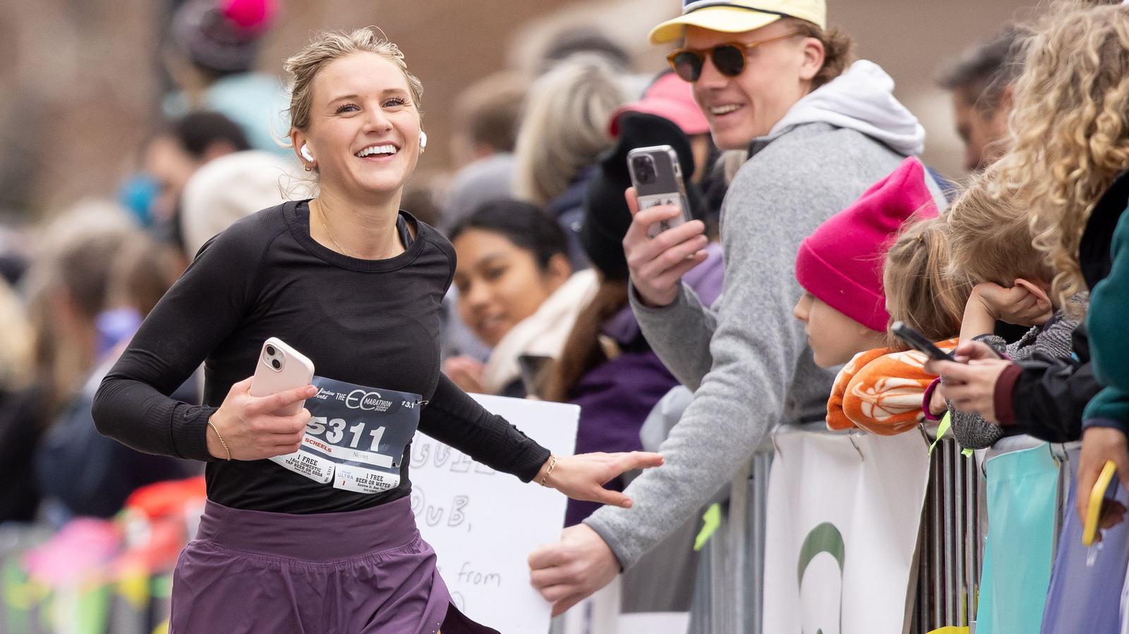 A smiling runner with bib number 5311 holds a phone while high-fiving a spectator. She's wearing a black top and purple shorts amidst a cheering crowd, including a man in sunglasses taking a photo.