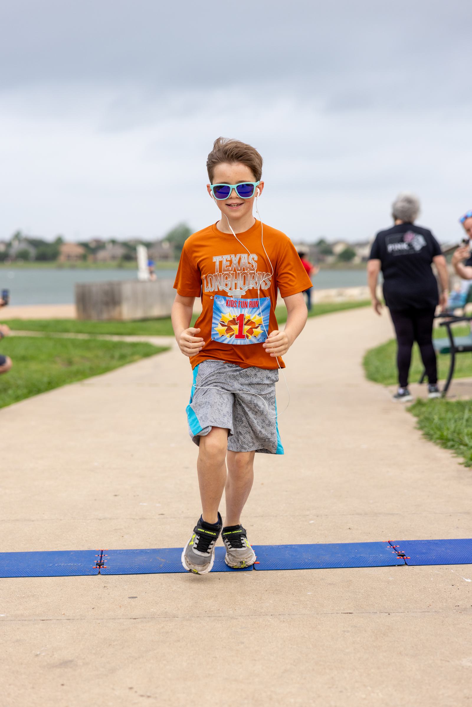 A young boy runs on a path beside a lake, wearing an orange shirt and blue sunglasses. He's listening to music with earphones. The cloudy sky and grassy area around the path are visible. Other people are in the background.