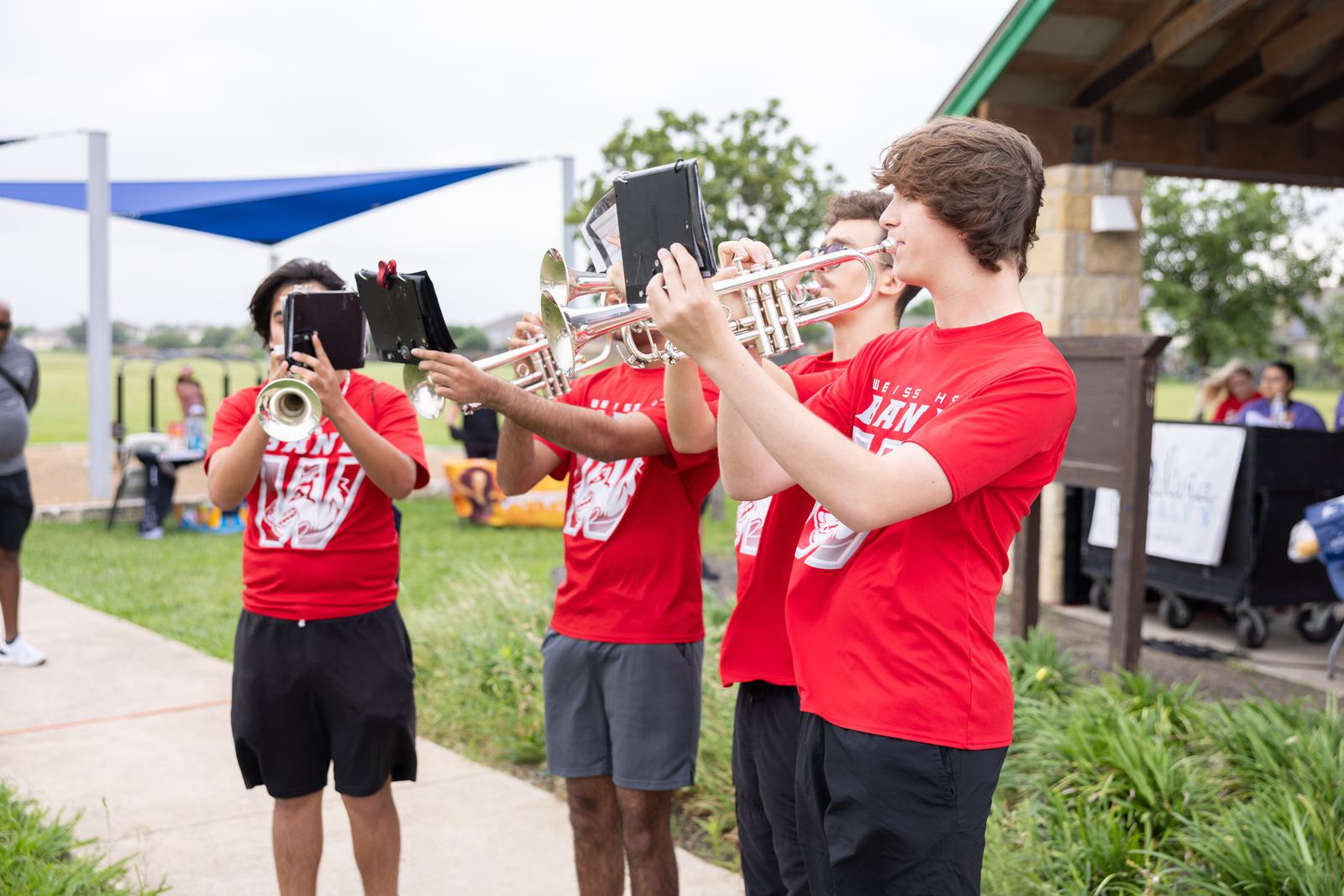 A group of four young people plays trumpets outdoors. They are wearing matching red shirts and black shorts. The background shows a park setting with grass, a covered area, and various outdoor structures.