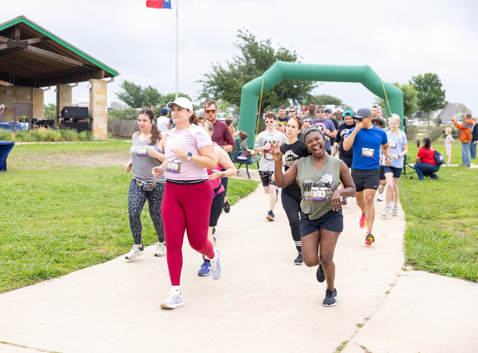 A group of diverse runners, wearing race bibs, jog along a paved path during an outdoor event. A green inflatable arch marks part of the course, and a pavilion with spectators is visible in the background. A flag flies above them in a cloudy sky.
