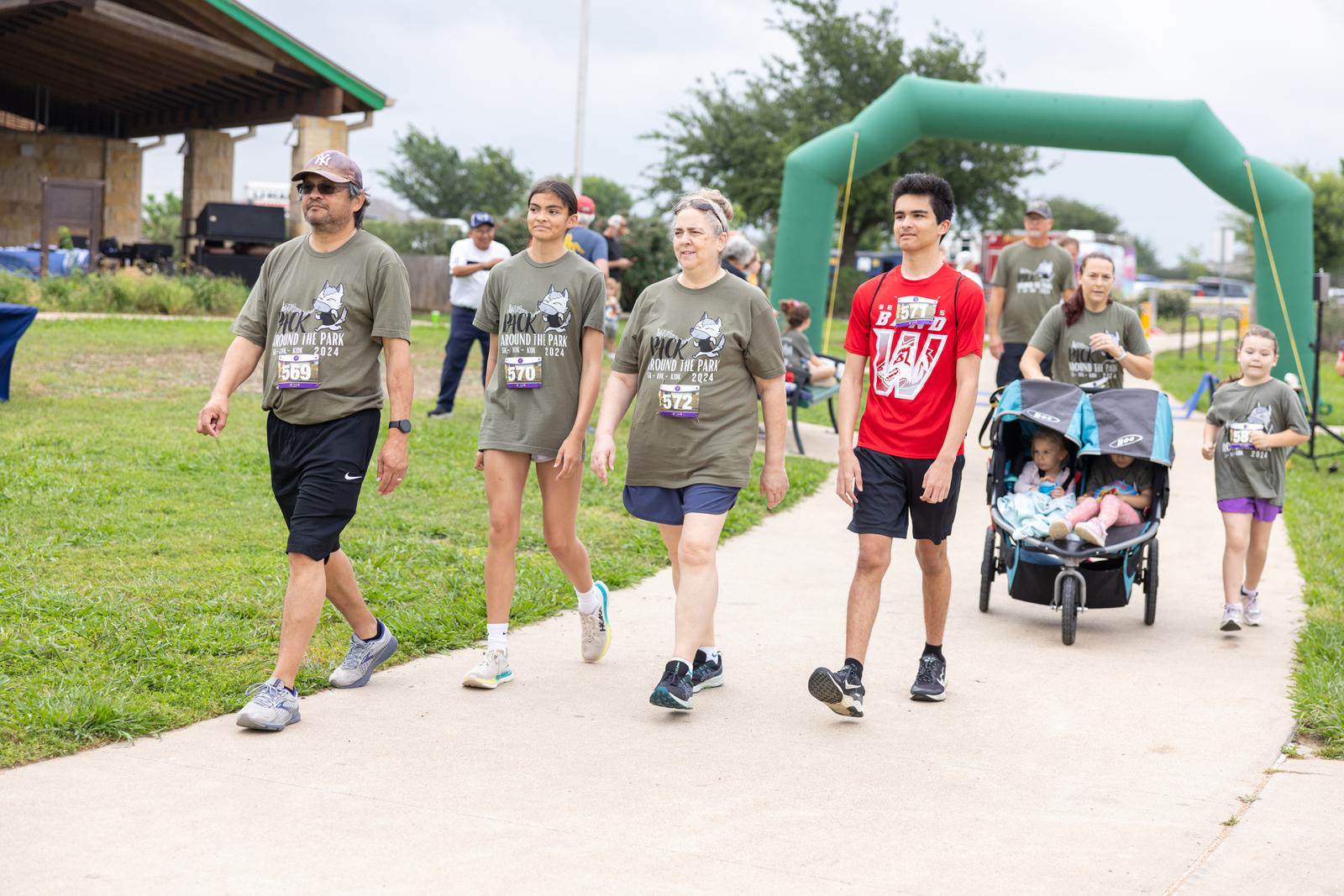 A group of people participating in a walkathon on a paved path. Some wear matching green shirts with event numbers, while one person wears a red shirt. A couple pushes a double stroller with two babies. An inflatable archway marks the path.