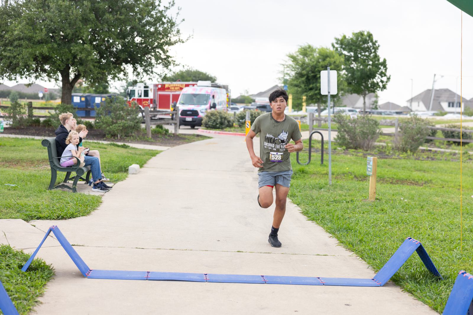 A boy in a green shirt and running shorts crosses a finish line on a park path. Two children sit on a bench in the background near emergency vehicles. The setting is outdoors with grass, trees, and overcast skies.