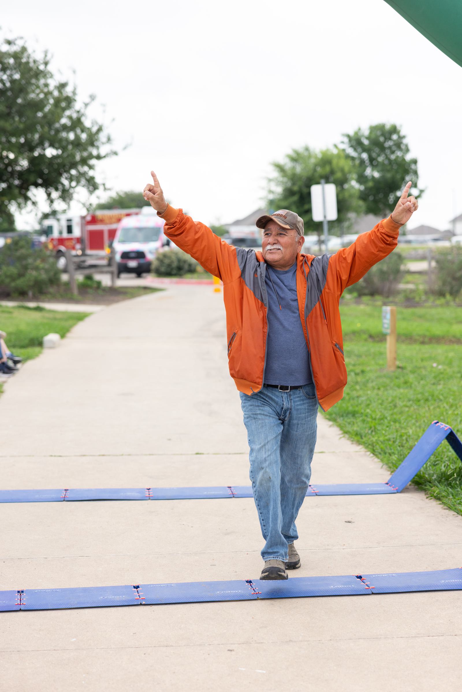 A man wearing a cap, orange jacket, and jeans raises his arms in celebration as he crosses a finish line on a park pathway. There are trees and a parked fire truck in the background.