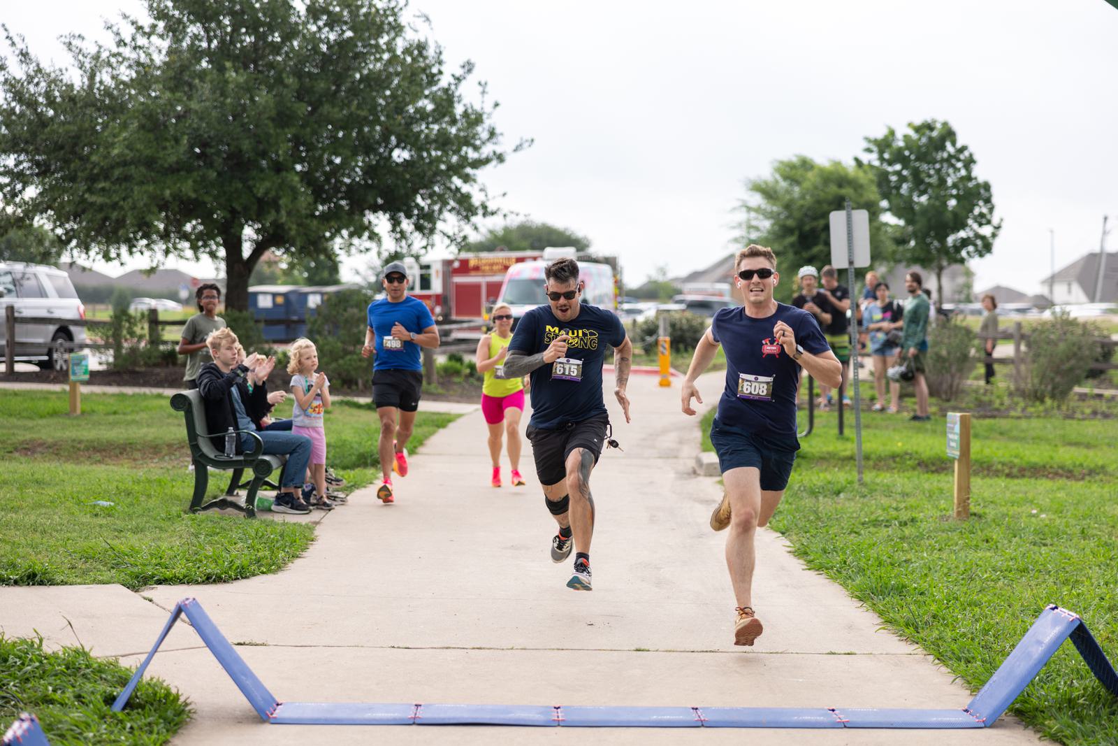 Participants sprint towards the finish line in a park race. Two men are leading, with others close behind. Spectators sit on a bench clapping, while others watch from the sidelines. Trees and a cloudy sky are in the background.