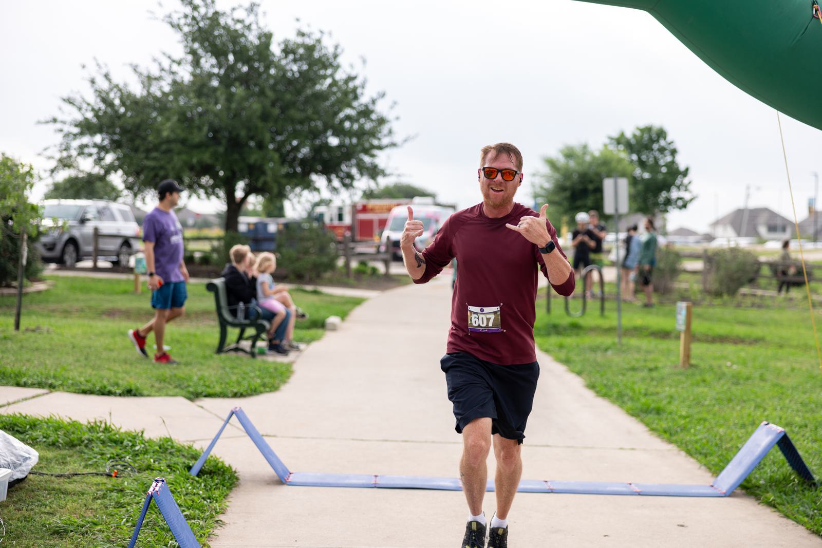 A man wearing sunglasses and a maroon shirt is running on a path, smiling and making a gesture with his hand. He holds a drink in his other hand. A few spectators sit on a bench nearby. Trees and grassy areas surround the path.