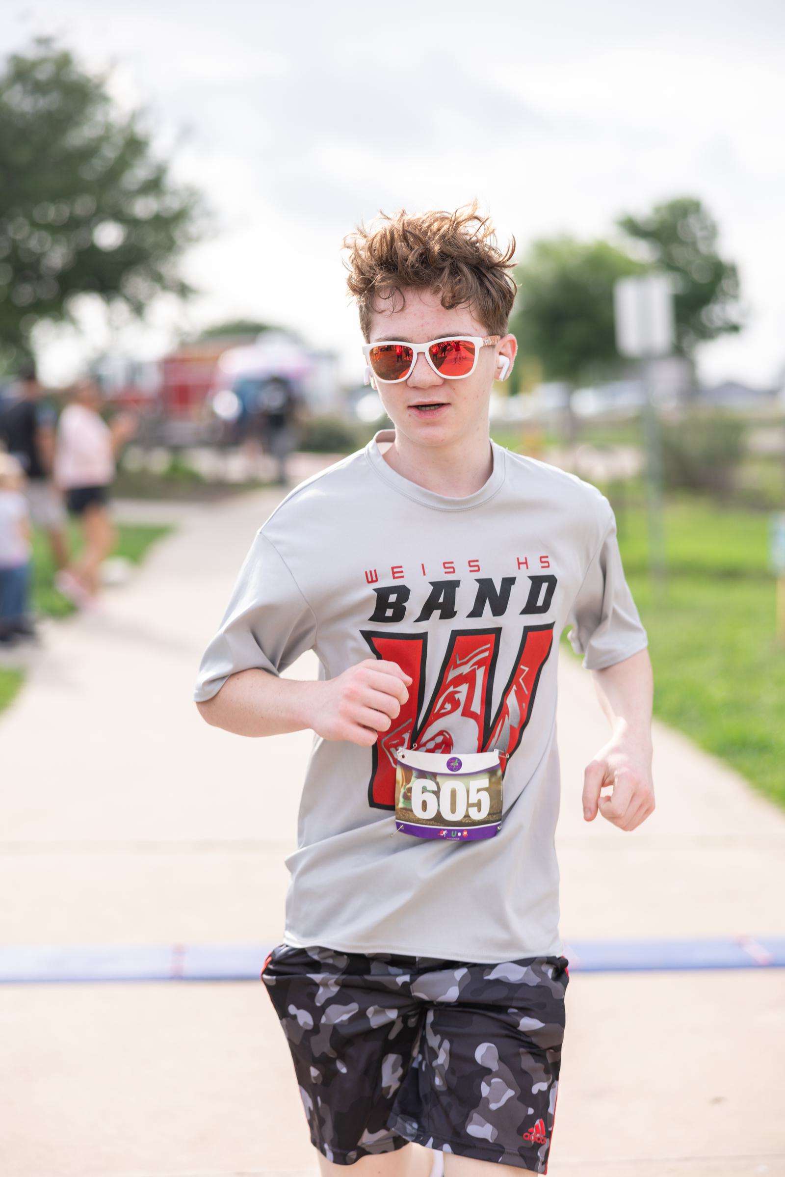 Young person running on a sidewalk in a race, wearing sunglasses and earbuds. They have a race bib numbered 605 and are dressed in a "Weiss HS Band" shirt and camo shorts. Trees and blurred spectators are in the background.