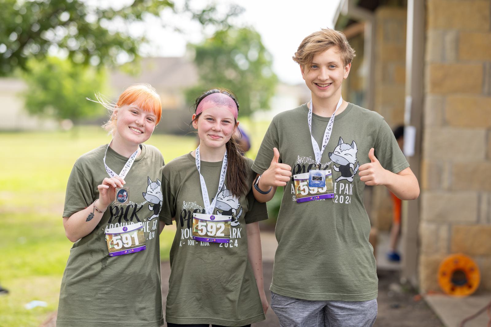 Three smiling people wearing green t-shirts and race bibs stand together outdoors, with two giving thumbs up and one holding a medal. They appear to be participating in a fun run event. A building and trees are visible in the background.