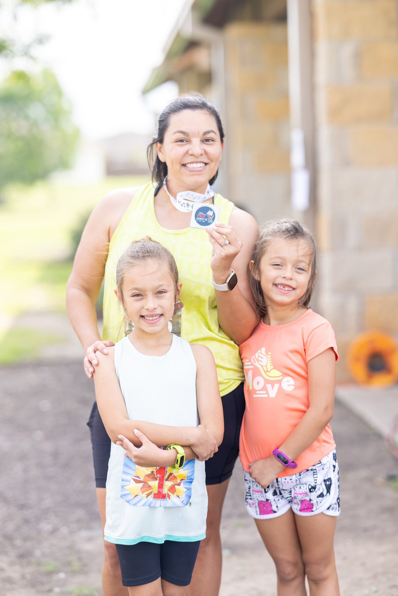 A woman smiling and holding a sticker stands outdoors with two young girls. The woman is in a yellow tank top, while the girls wear light blue and orange tops. They are in front of a stone wall on a sunny day.