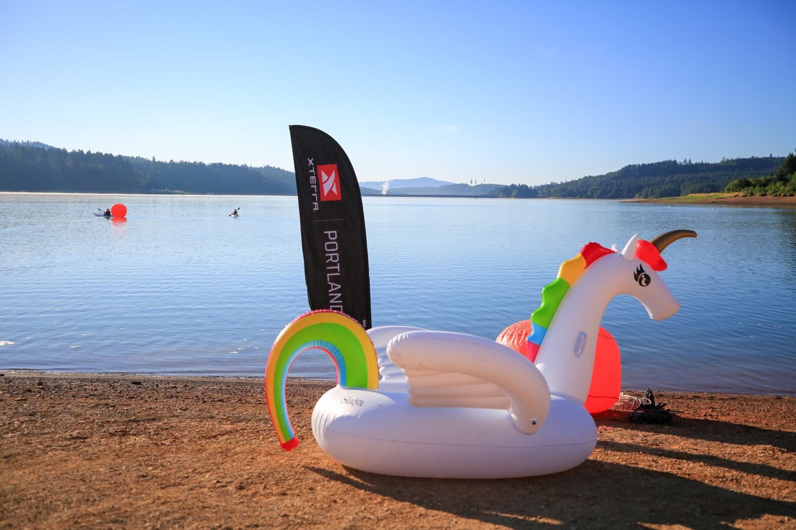 A large inflatable unicorn float rests on the sandy shore of a calm lake, with a dark promotional banner and red buoys in the background. Forested hills line the horizon under a clear blue sky.