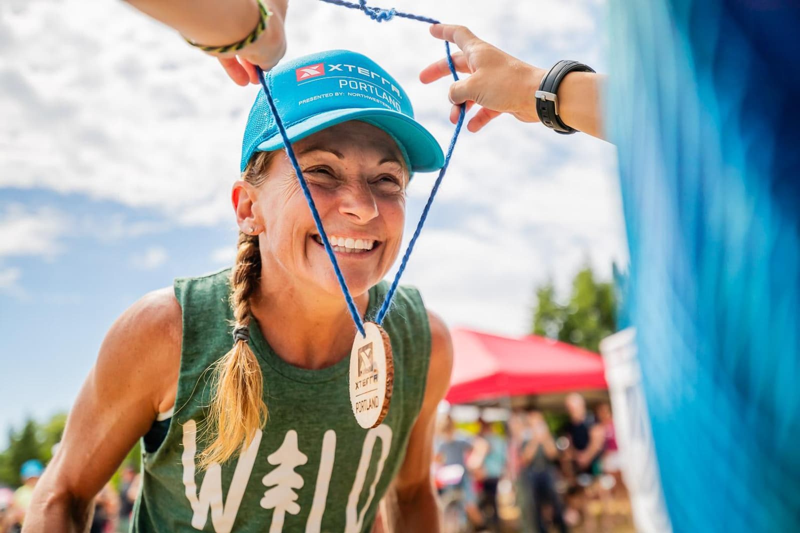 A woman in a green tank top and blue cap smiles as she receives a medal on a blue ribbon. The background shows a sunny outdoor setting with a red tent and blurred spectators.