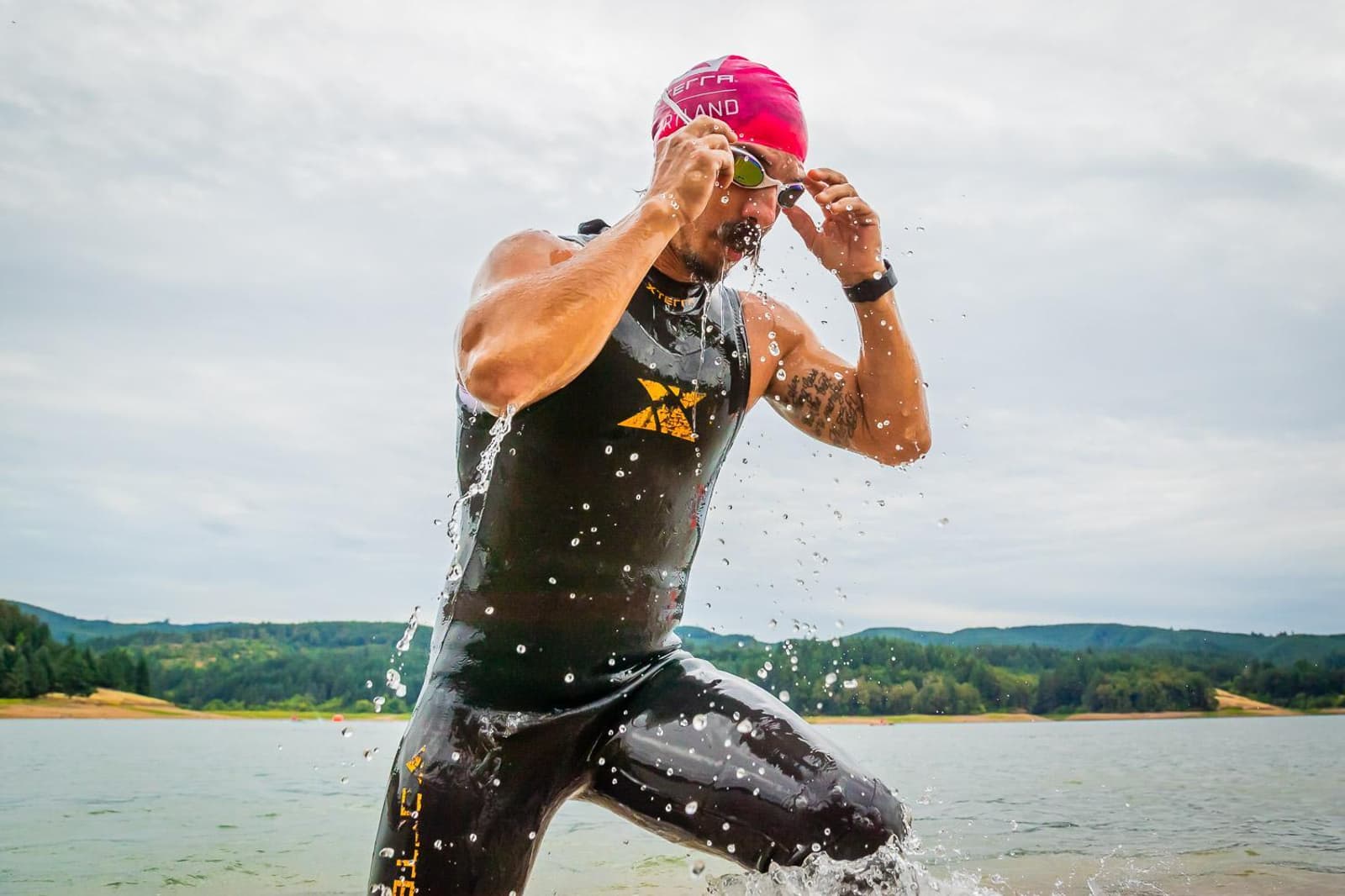 A triathlete emerges from the water, adjusting his goggles. He's wearing a black wetsuit and a pink swim cap. Water droplets splash around him, with a scenic backdrop of a lake and distant green hills under a cloudy sky.