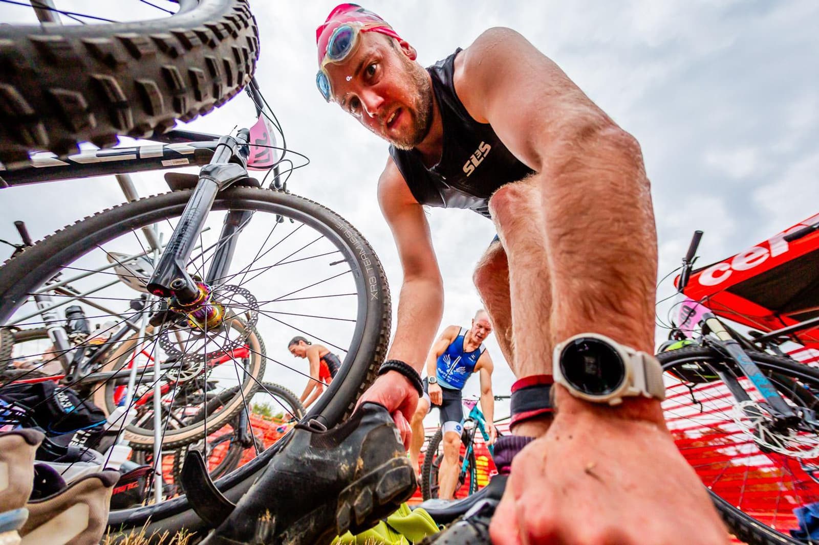 A triathlete in a black suit, wearing a swim cap and goggles, prepares to transition between events. He leans down, adjusting his gear beside several bicycles. Other athletes and equipment are visible in the background.