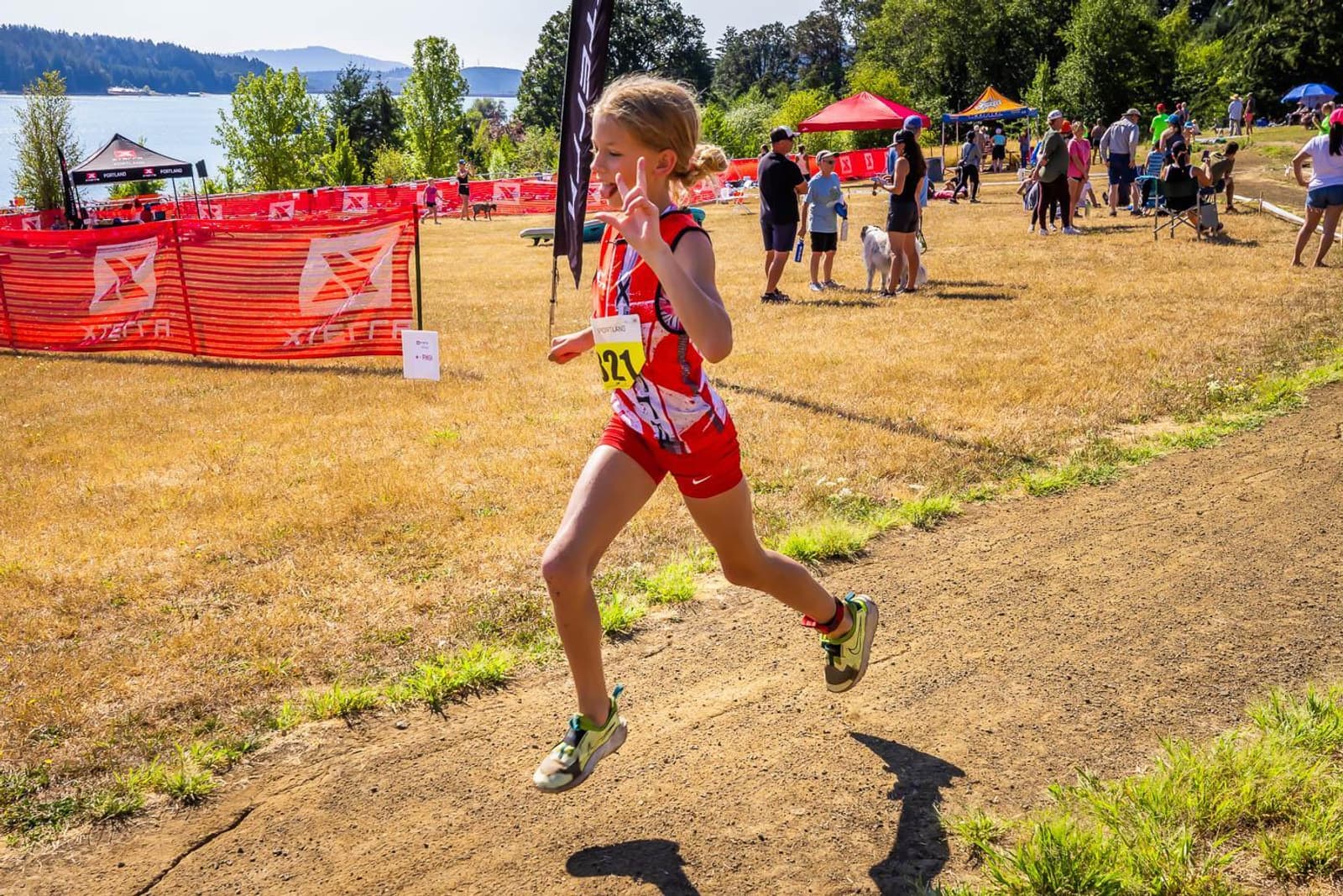 Young athlete in red and white gear running on a dirt path during an outdoor event. Spectators, tents, and trees are visible in the background under a clear blue sky.