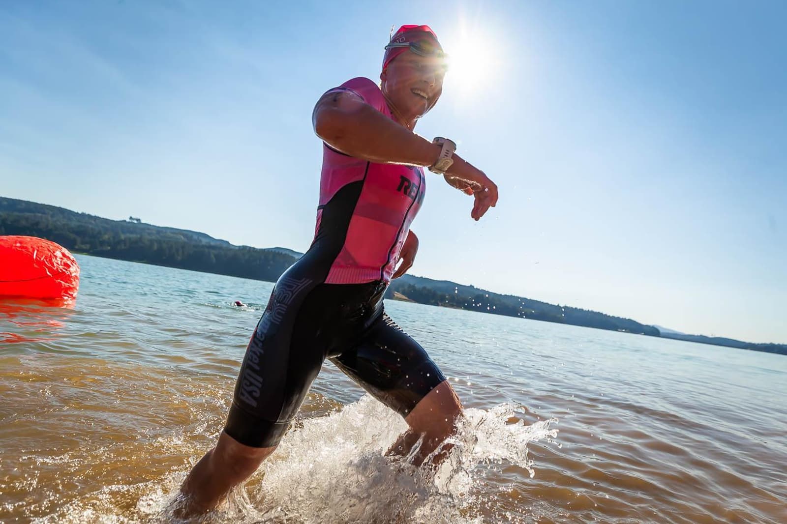 A person in a pink and black triathlon suit emerges from a lake, splashing water. The sun shines brightly behind them, creating a vivid glare. A large red buoy is visible on the water, with hills in the distant background.