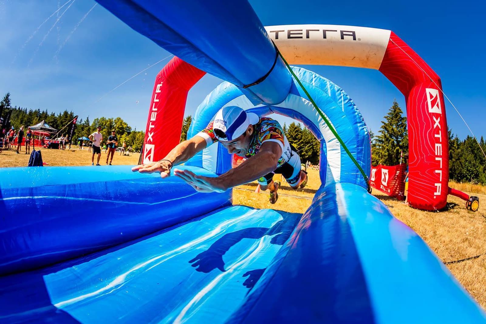 A person in athletic gear dives onto a slip-and-slide during an outdoor event. The slide is blue and part of an obstacle course with an inflatable arch marked "XTERRA." Spectators and tents are visible in the background under a clear blue sky.