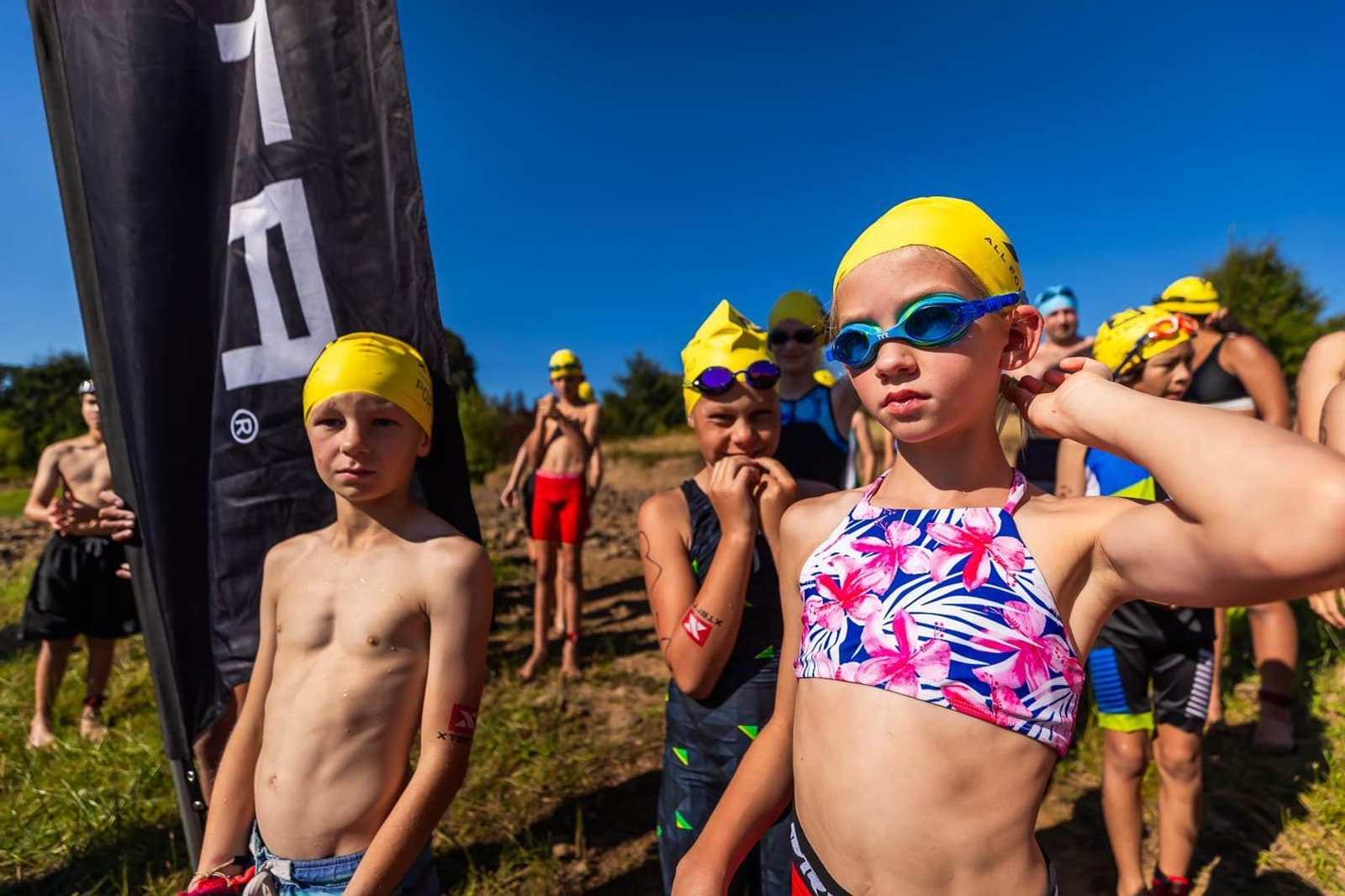 Children prepare for a race, wearing yellow swim caps and swimwear. A girl adjusts her goggles while others stand nearby under a clear blue sky, with a black flag in the background. The atmosphere is lively and focused.