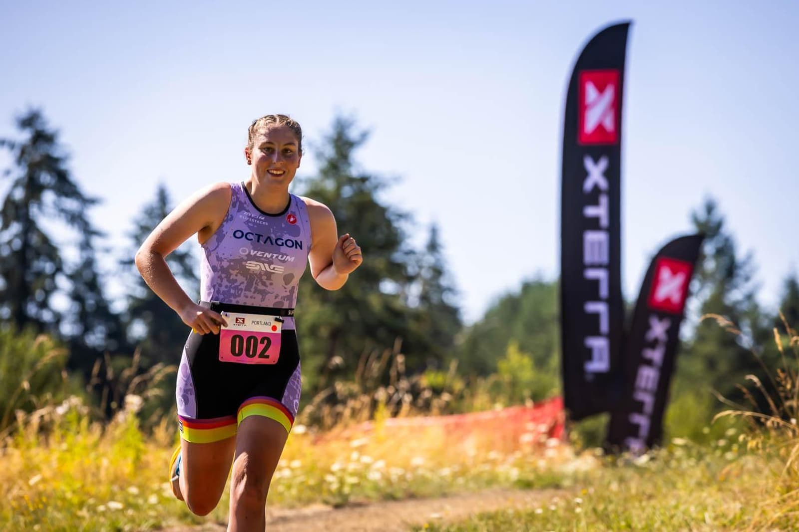 A person wearing a race bib number 002 is running on a dirt trail during an outdoor race. They're dressed in athletic gear with a purple top. Two banners with the XTERRA logo stand in the background amidst trees and grass on a sunny day.