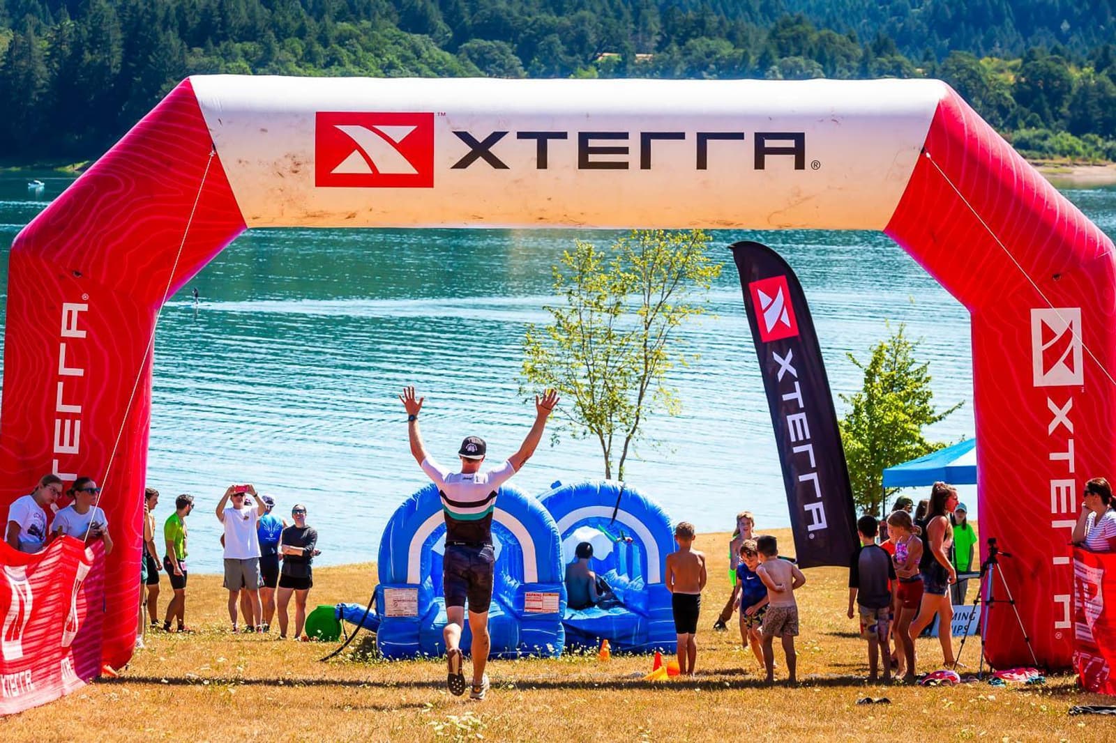 A person raises their arms in victory while crossing the XTERRA finish line beside a lake. Spectators and participants stand nearby, with wooded hills in the background under a clear sky.