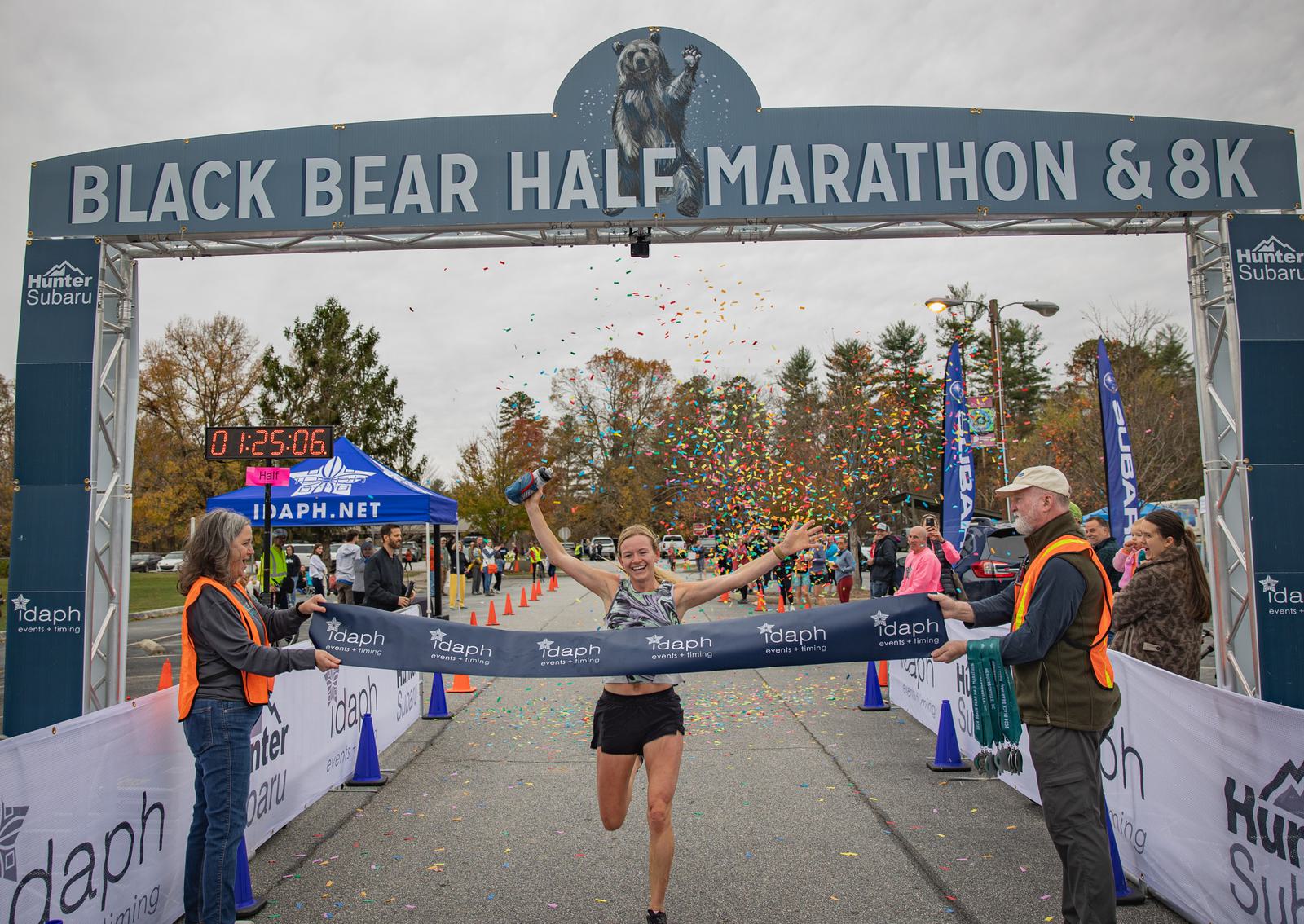 A runner joyfully crosses the finish line at the Black Bear Half Marathon & 8K, arms raised in victory as confetti falls. Spectators and officials stand nearby, and a race clock shows the finishing time.