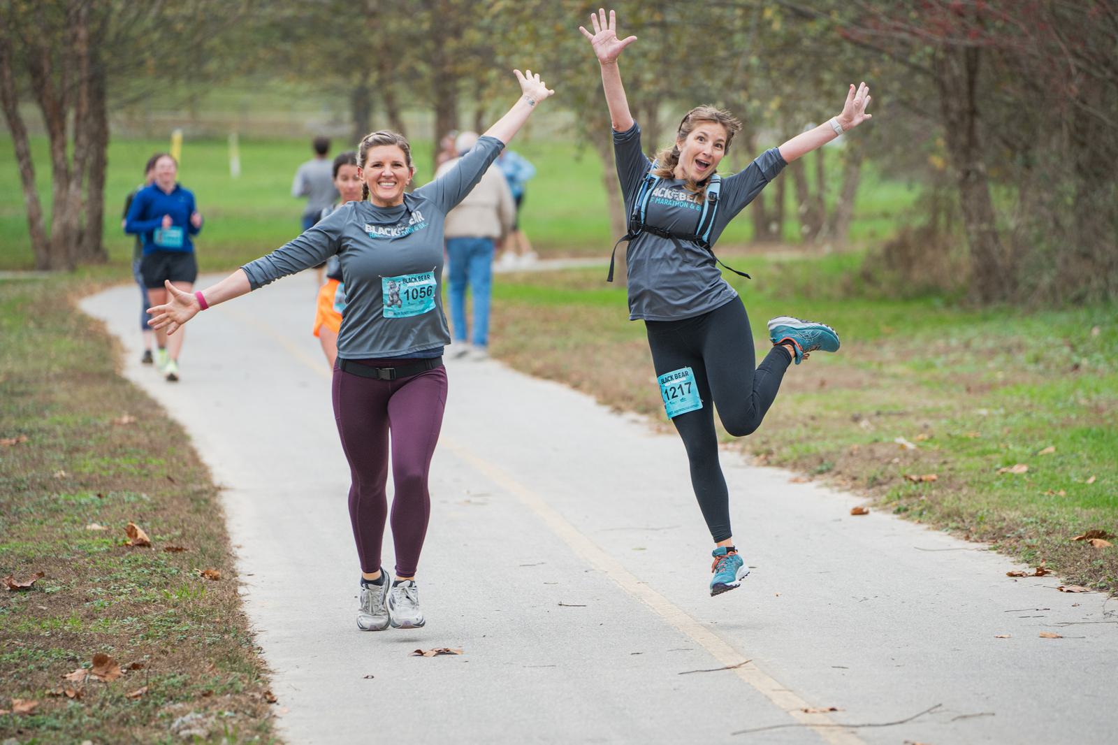 Two women in athletic wear joyfully run on a paved park trail, smiling with arms raised; one woman is mid-jump. Other runners are visible in the background among trees and grass.