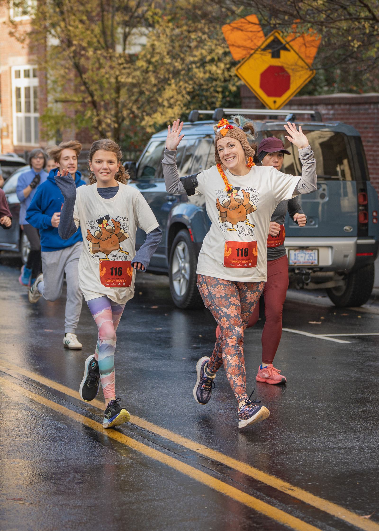 Two smiling runners, a woman and a boy, wave as they run down a wet street during a race. They wear matching event T-shirts with cartoon turkeys and colorful leggings. Other participants and parked cars are visible in the background.