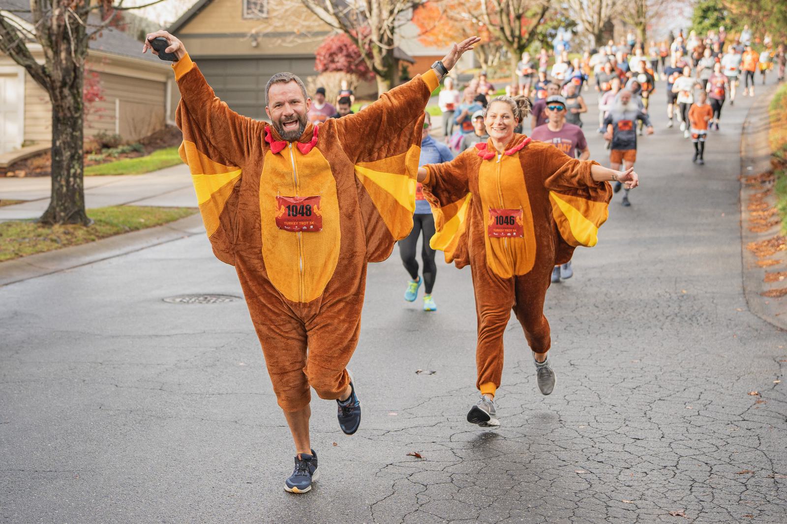 Two people in turkey costumes joyfully run toward the camera during a race, arms raised and smiling, with many other runners and autumn trees visible in the background.