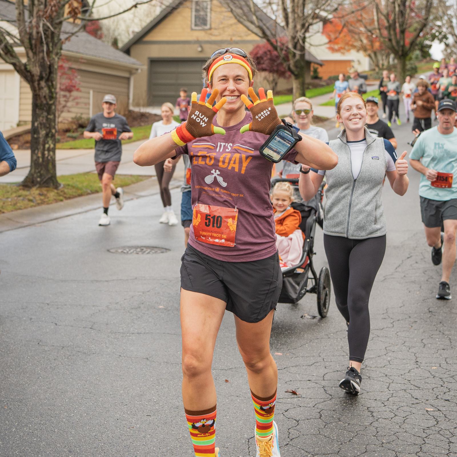 A smiling runner wearing a "It's All Day" shirt, colorful socks, and "RUN HAPPY" gloves raises her hands for the camera during a race. Other runners and a stroller are visible behind her on a wet neighborhood street.