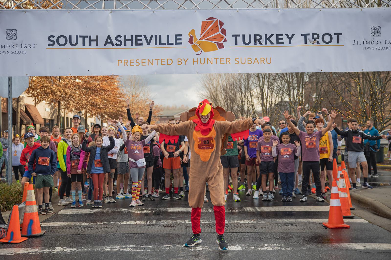A person in a turkey costume stands in front of a crowd of runners at the start line of the South Asheville Turkey Trot, with the event banner overhead and autumn trees in the background.