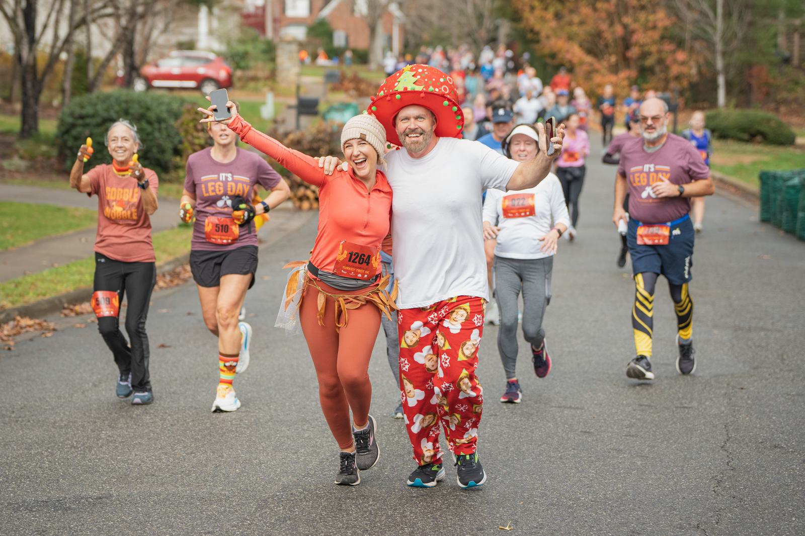 A smiling man in festive red pajamas and a large red hat runs alongside a woman in orange athletic wear, both with arms raised, leading a group of happy runners in a race on a tree-lined street.