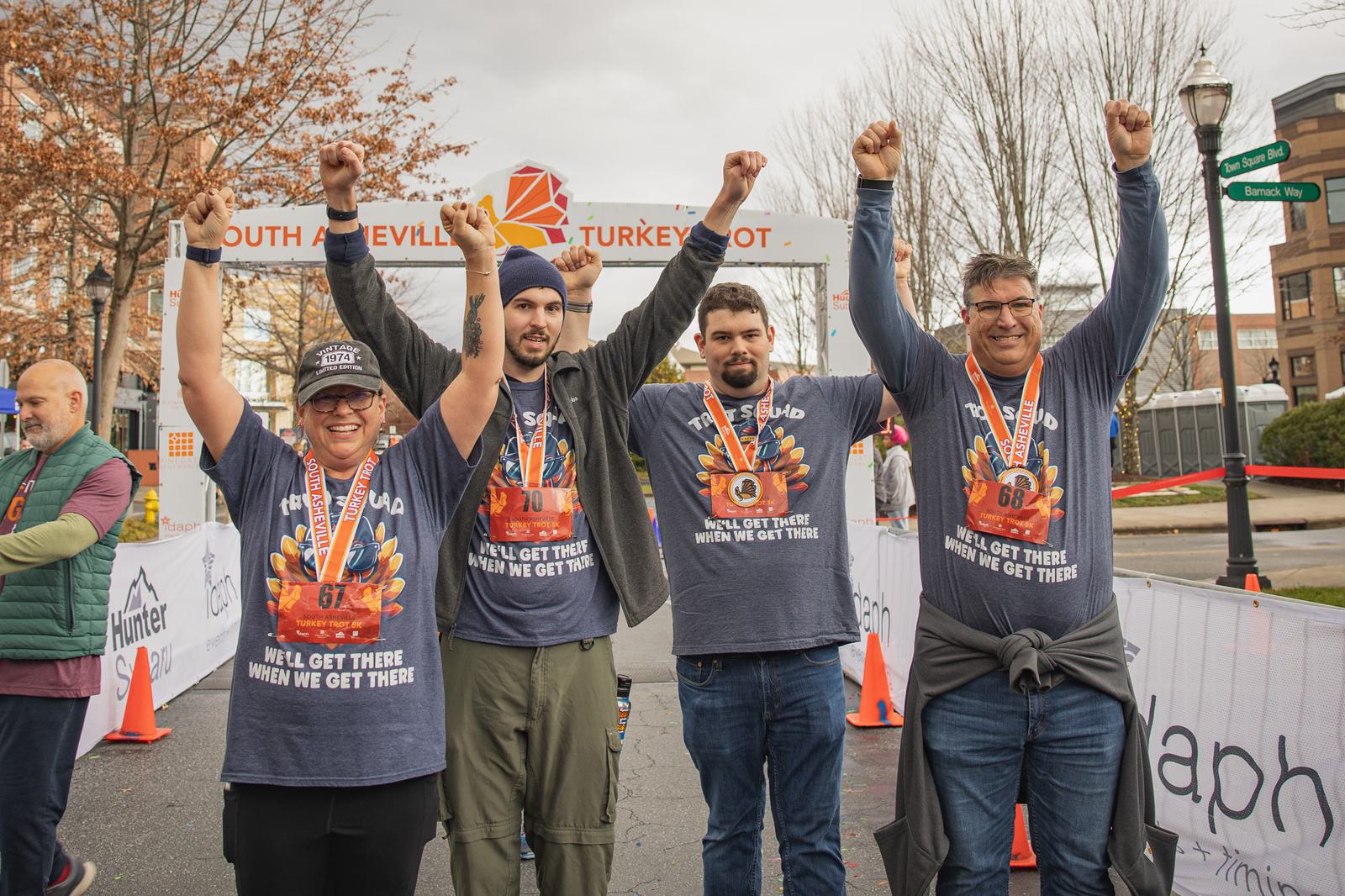 Four smiling runners wearing medals raise their arms in celebration at a finish line. They're dressed in matching shirts, standing on a city street with event banners and autumn trees in the background.