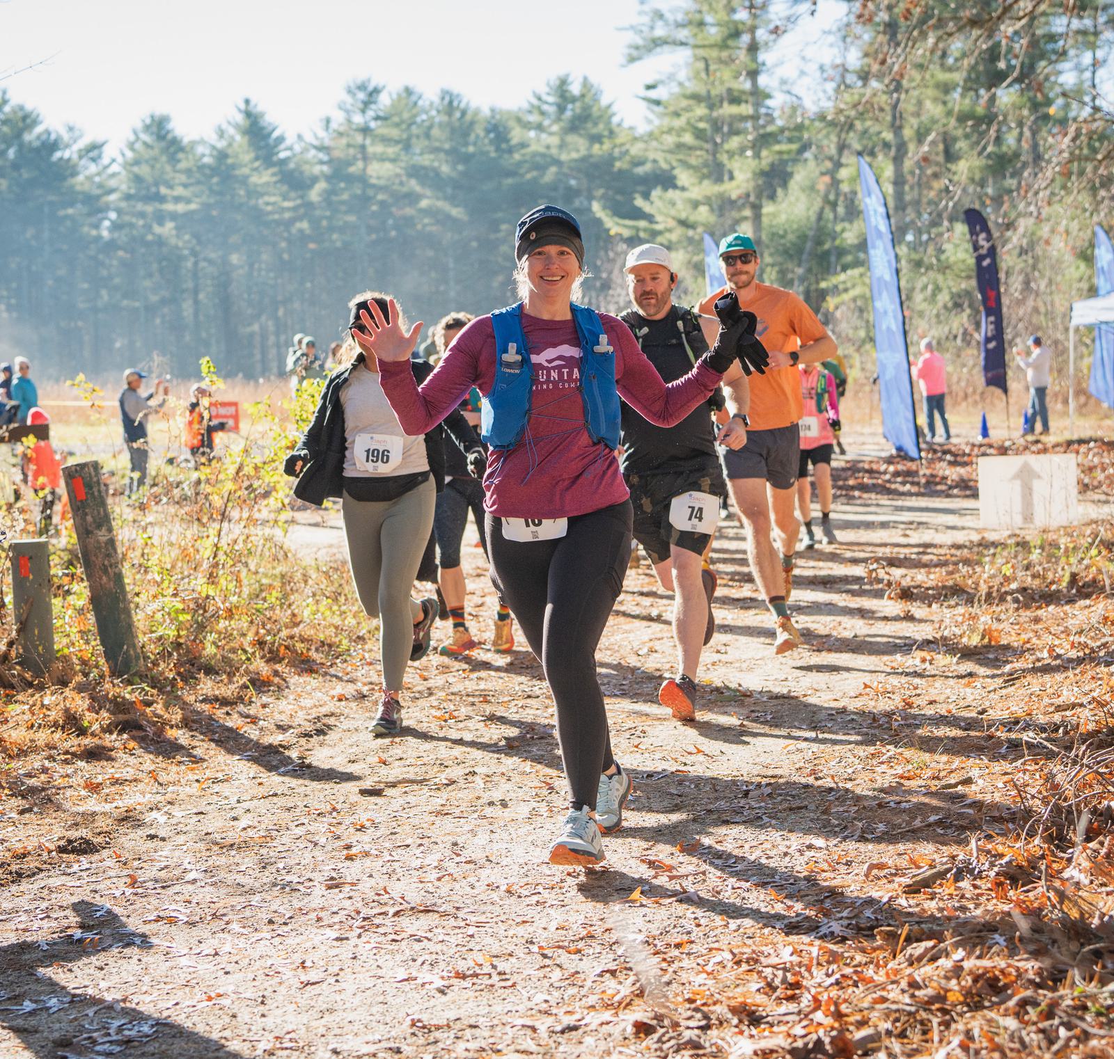 A group of runners participate in a trail race through a sunlit forest. The leading woman smiles and waves at the camera, while others follow behind, all wearing race bibs and athletic gear.