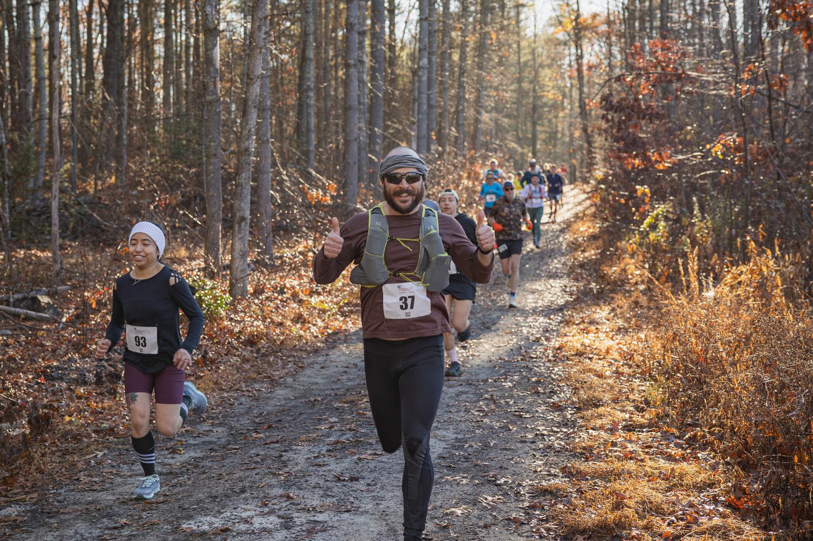 A group of runners participates in a trail race through a sunlit autumn forest. The man in front, wearing a race bib numbered 37, smiles and gives thumbs up while others follow behind him on the dirt path.
