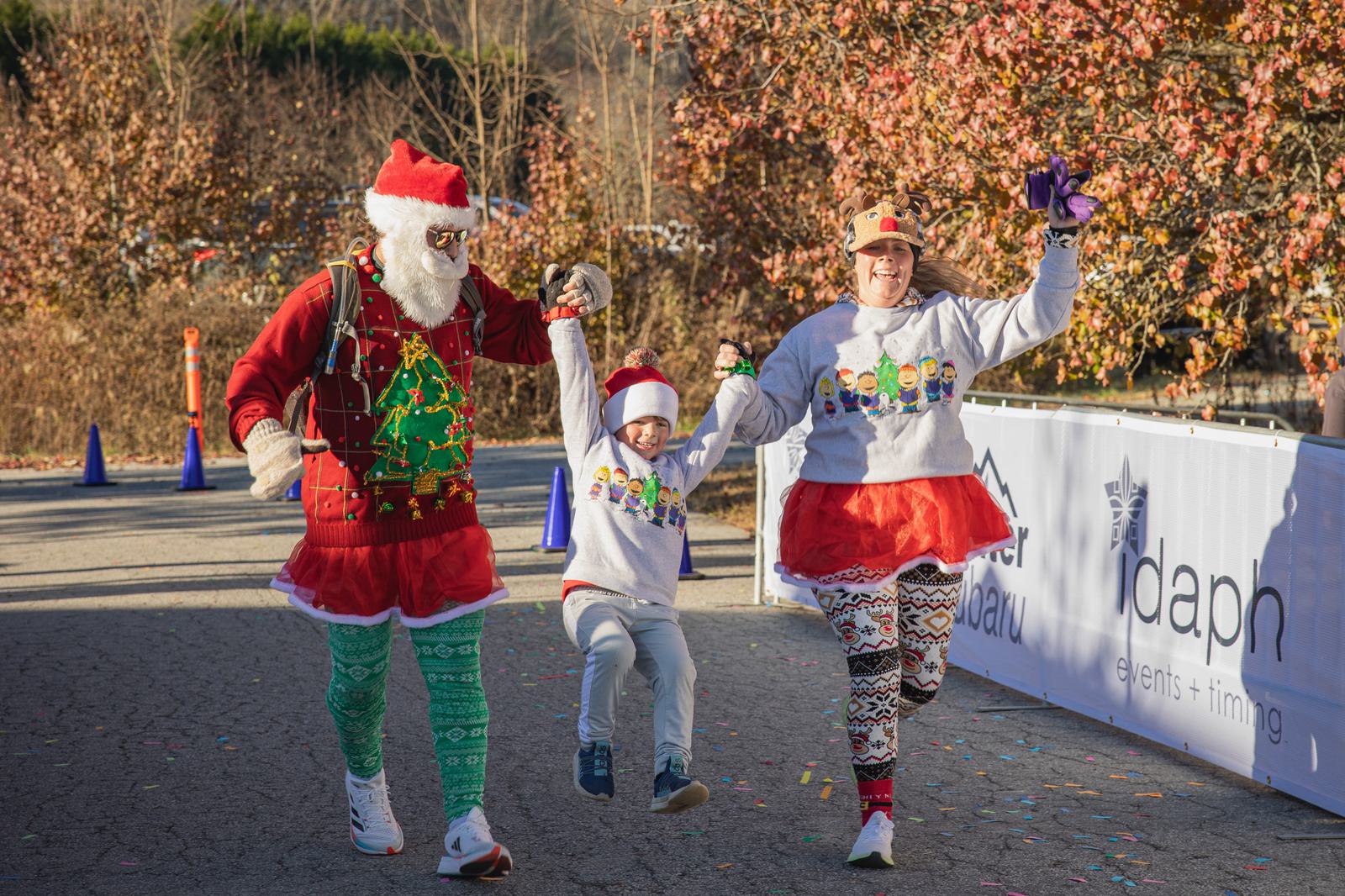 Three people in festive holiday outfits, including Santa hats and Christmas sweaters, hold hands and smile as they cross a finish line together. The child in the center is being lifted off the ground. Autumn trees are in the background.