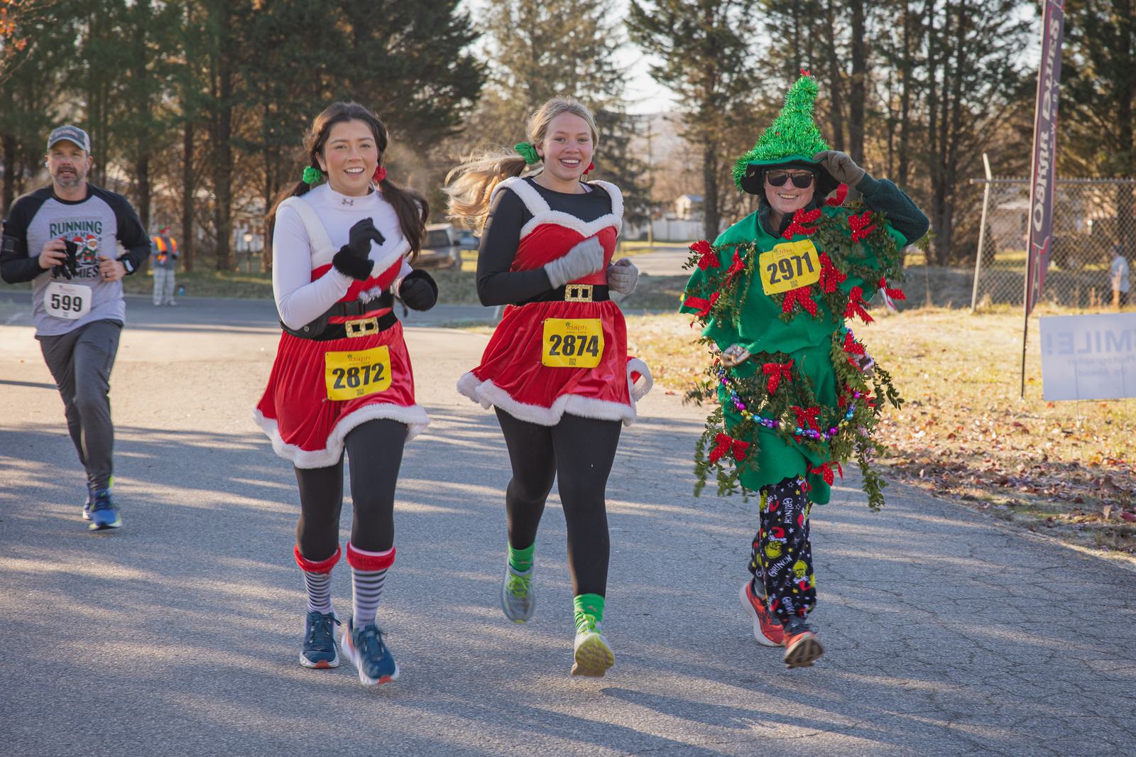 Three people in festive costumes—two dressed as Mrs. Claus and one as a Christmas tree—run in a race, smiling and wearing numbered bibs. Other runners and trees are visible in the background.