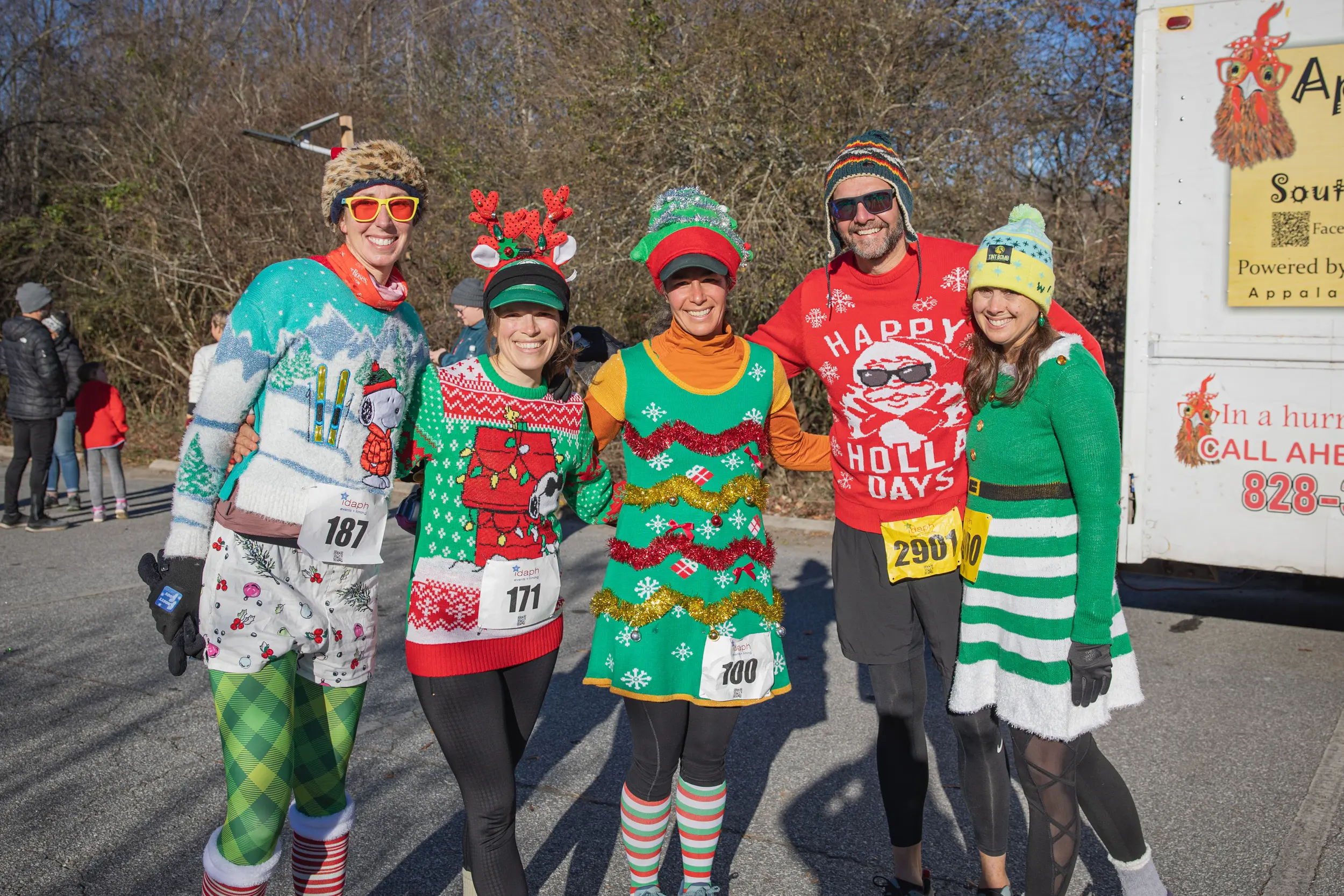 Five people in festive holiday costumes and race bibs smile together outdoors during a sunny day. They wear Christmas sweaters, hats, and accessories, standing on a paved path near trees and a food truck.