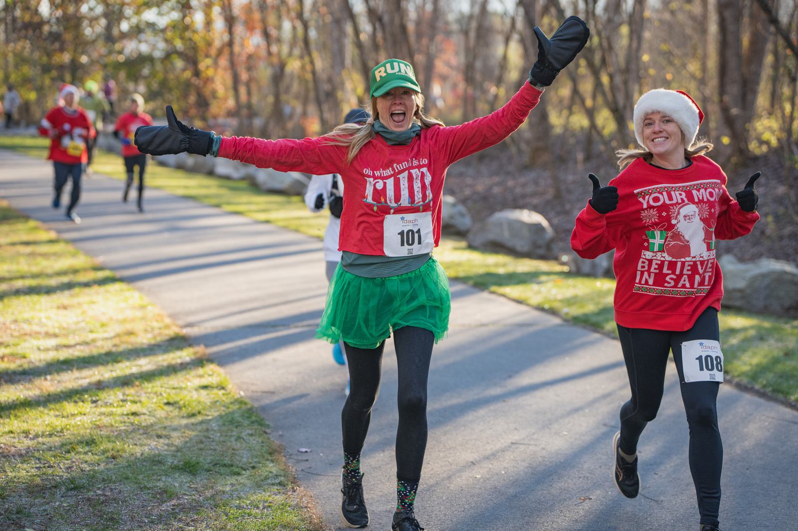 Two women in festive holiday outfits, wearing numbered race bibs, smile and gesture playfully as they run on a paved park trail during a winter fun run, surrounded by trees and other participants.
