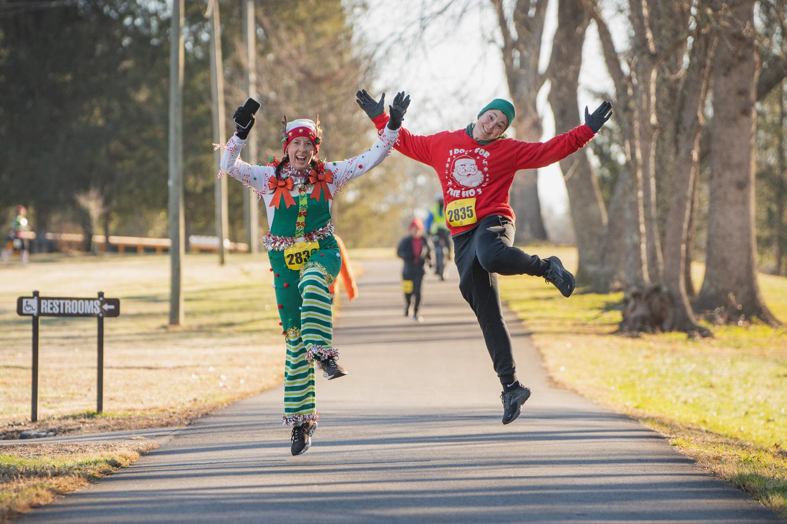Two people dressed in festive holiday outfits and race bibs jump joyfully in the air on a park path during a fun run. There are trees, sunlight, and a restroom sign visible in the background.