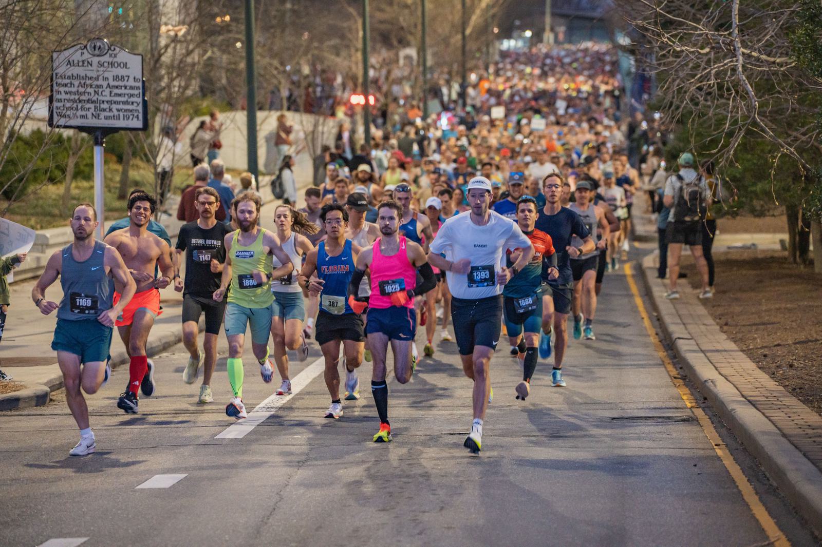 A large group of runners, wearing colorful athletic gear, races down a city street during a marathon or running event, with spectators and trees lining the road.