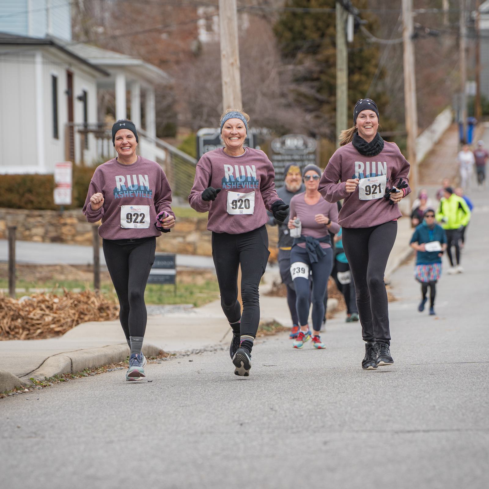 Three women wearing matching "Run Asheville" sweatshirts and race bibs (922, 920, 921) jog and smile during a road race, with other runners and houses visible in the background.