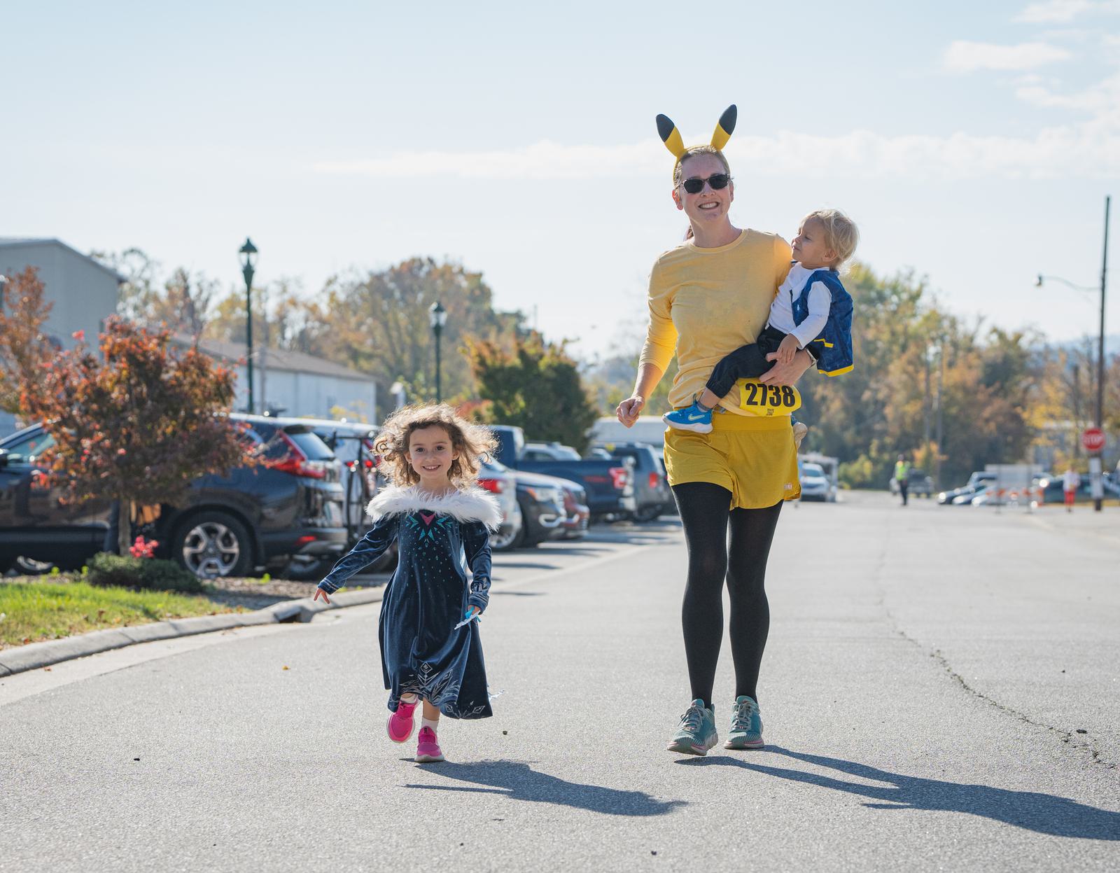 An adult in a yellow Pikachu costume runs on a street holding a small child, while a young girl in a blue dress and pink shoes runs ahead, all smiling on a sunny day.